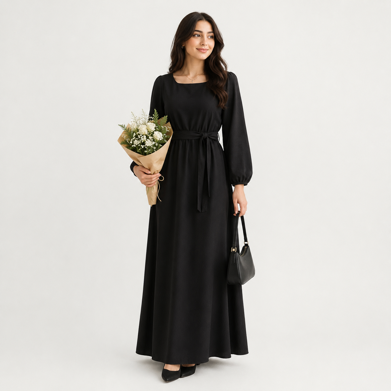 A woman modeling a long black maxi dress with long sleeves, a square neckline, and a self-tie waist belt, holding a bouquet of white flowers and a black handbag against a light gray studio background.