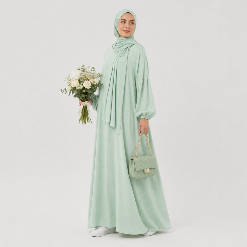 A woman wearing a light sage green hijab, long-sleeve maxi abaya, and matching handbag, holding a bouquet of white flowers against a plain light gray studio background.