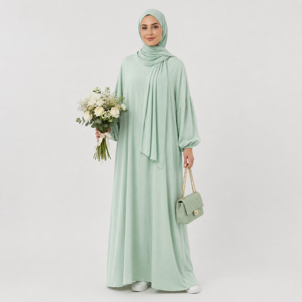 A woman wearing a light sage green hijab, long-sleeve maxi abaya, and matching handbag, holding a bouquet of white flowers against a plain light gray studio background.