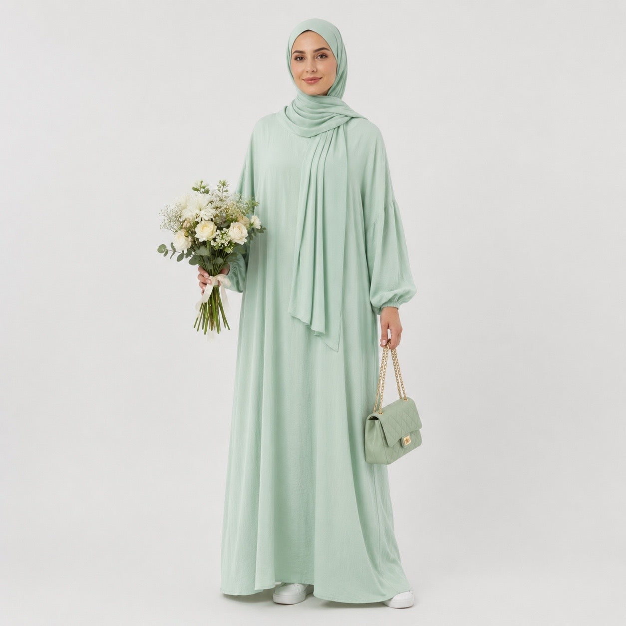 A woman wearing a light sage green hijab, long-sleeve maxi abaya, and matching handbag, holding a bouquet of white flowers against a plain light gray studio background.