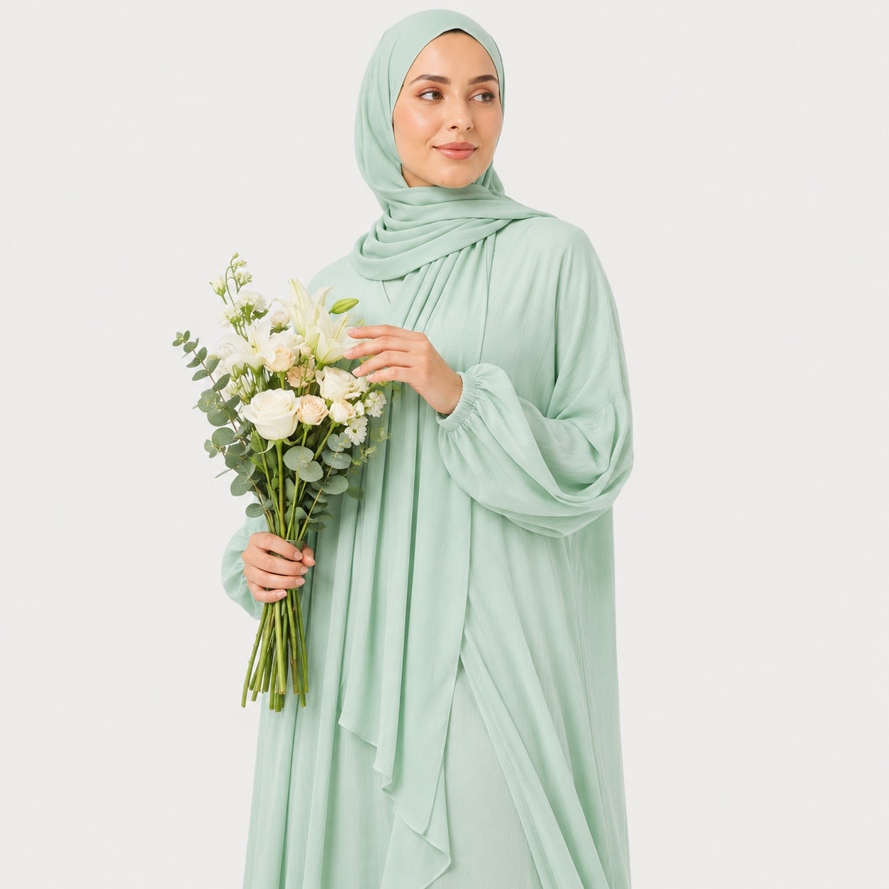 A woman wearing a light sage green hijab, long-sleeve maxi abaya, and matching handbag, holding a bouquet of white flowers against a plain light gray studio background.