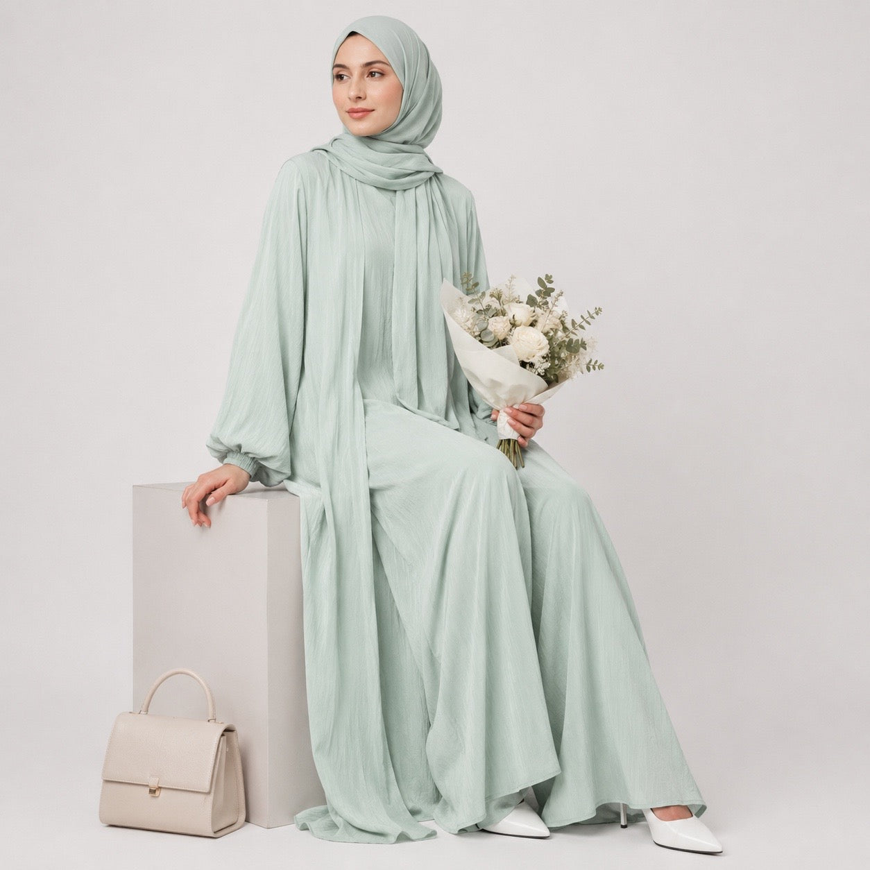 A woman wearing a light sage green hijab, long-sleeve maxi abaya, and matching handbag, holding a bouquet of white flowers against a plain light gray studio background.