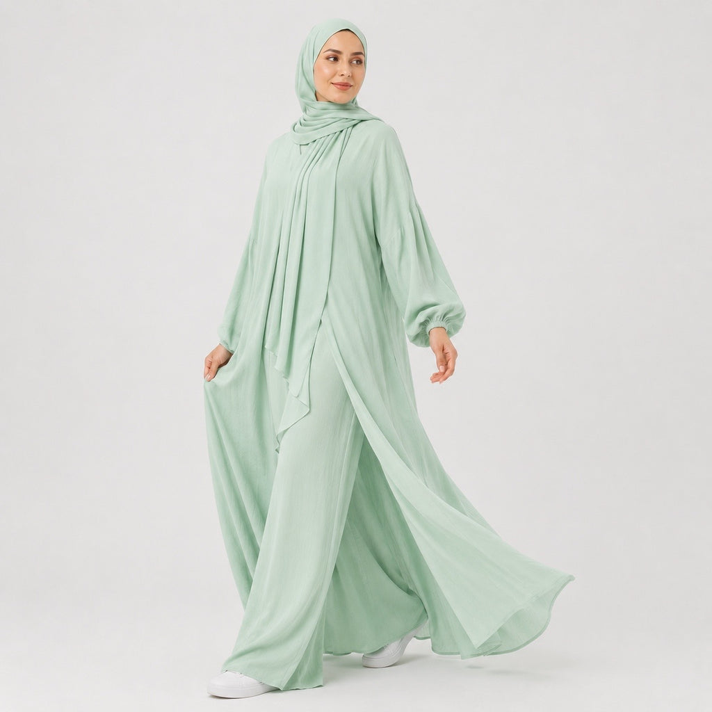 A woman wearing a flowing sage green modest outfit with a matching hijab, wide-leg pants, and a long draped top, photographed against a clean light gray studio background.