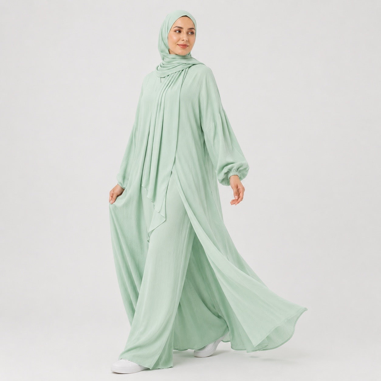 A woman wearing a flowing sage green modest outfit with a matching hijab, wide-leg pants, and a long draped top, photographed against a clean light gray studio background.