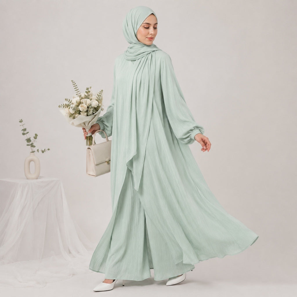 A woman wearing a light sage green hijab, long-sleeve maxi abaya, and matching handbag, holding a bouquet of white flowers against a plain light gray studio background.