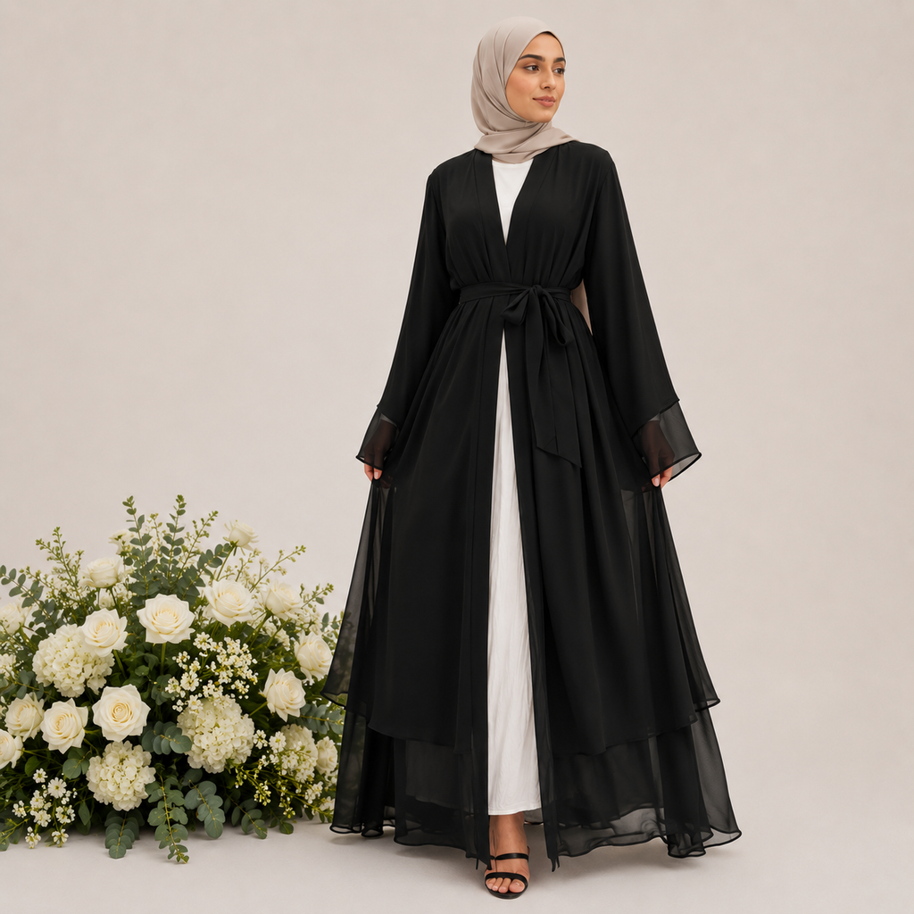 Woman wearing a black abaya with a white dress underneath, standing next to a floral arrangement on a light background.