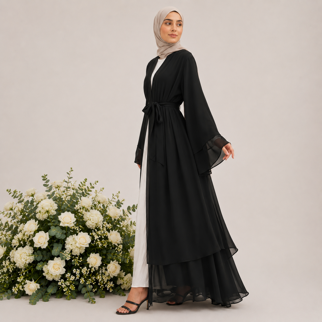 Woman wearing a black abaya with sheer sleeves standing next to white flowers on a light gray background