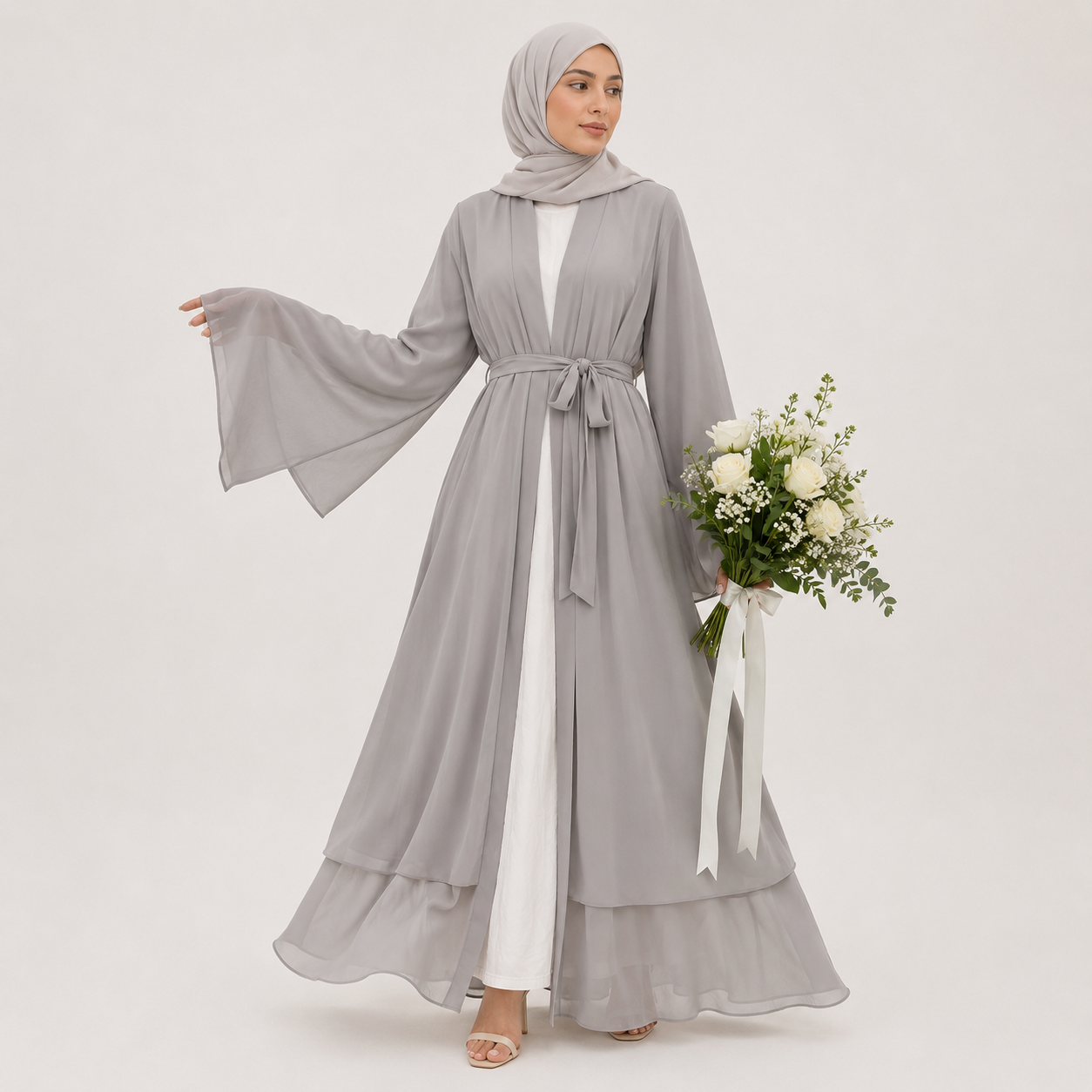 Woman wearing a gray abaya with a white dress underneath, holding a bouquet of flowers on a light gray background.