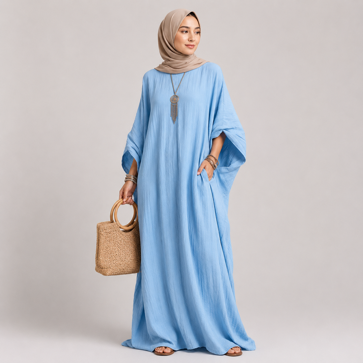 Woman wearing a light blue dress and beige hijab holding a woven bag on a plain background