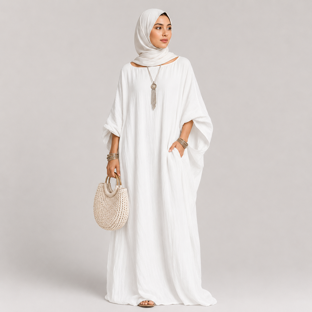 Woman wearing a white dress and hijab holding a woven bag on a plain background