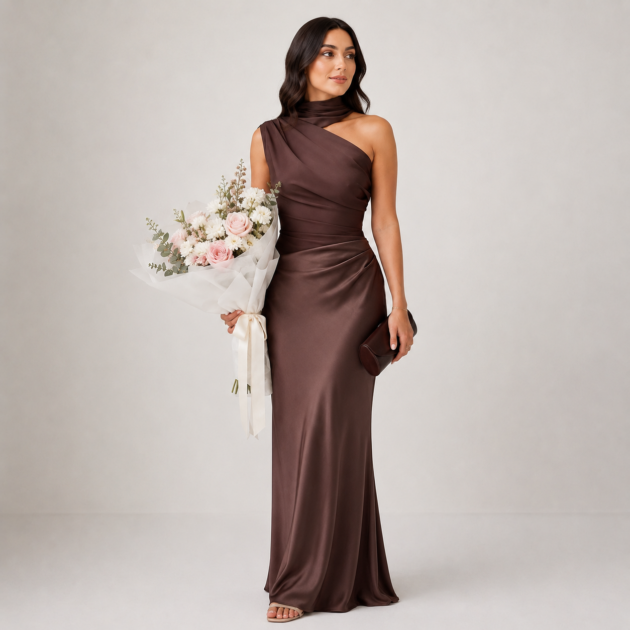 Woman in a brown one-shoulder dress holding flowers and a clutch against a plain background