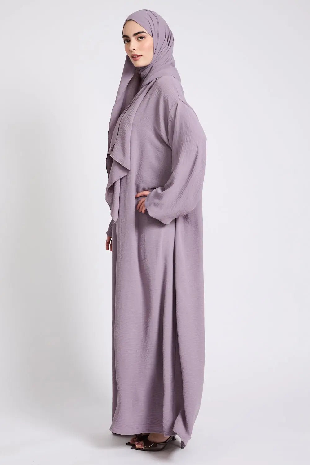 Woman wearing a long, light purple abaya with a matching hijab on a white background