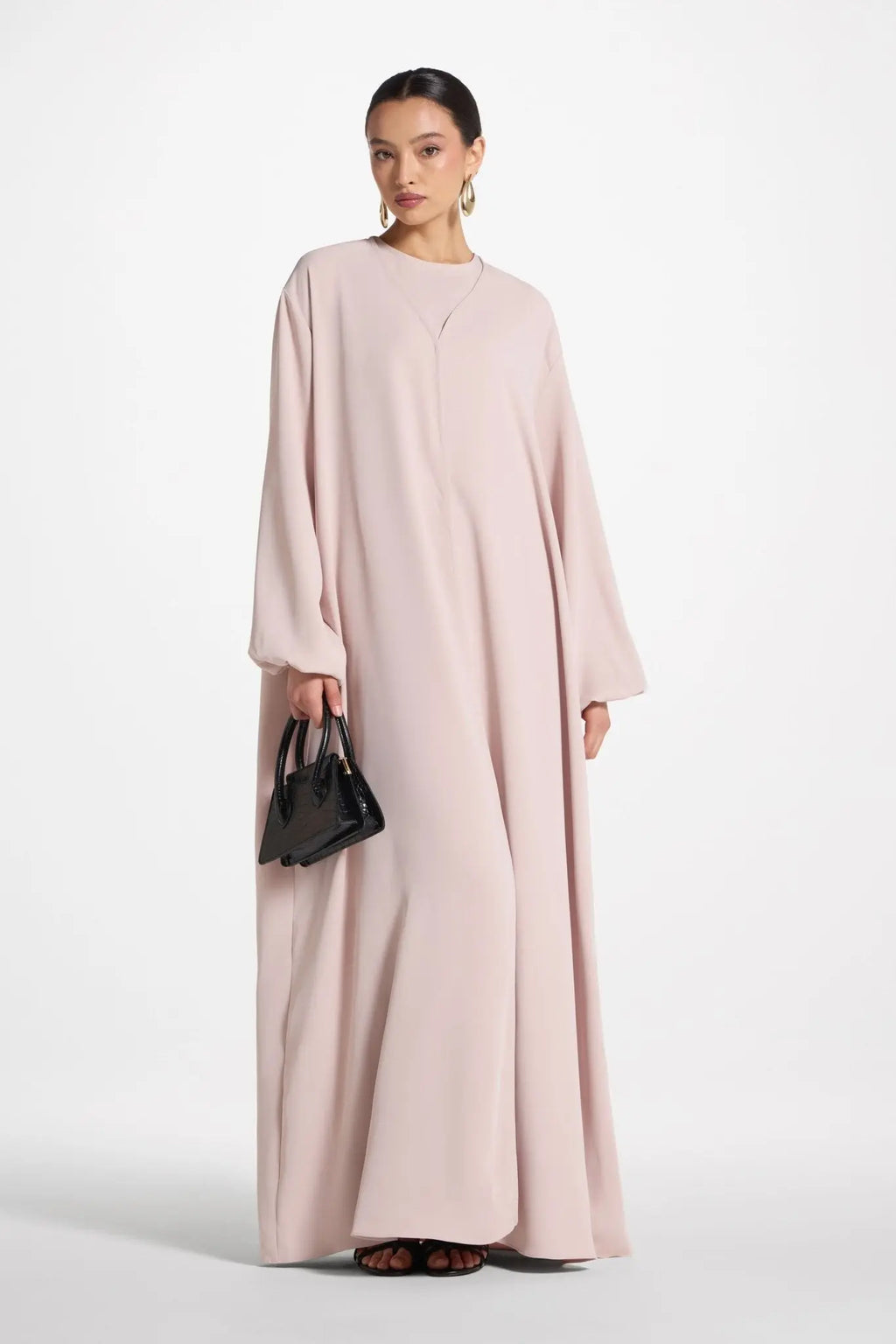 Woman wearing a long light pink dress holding a black handbag on a white background