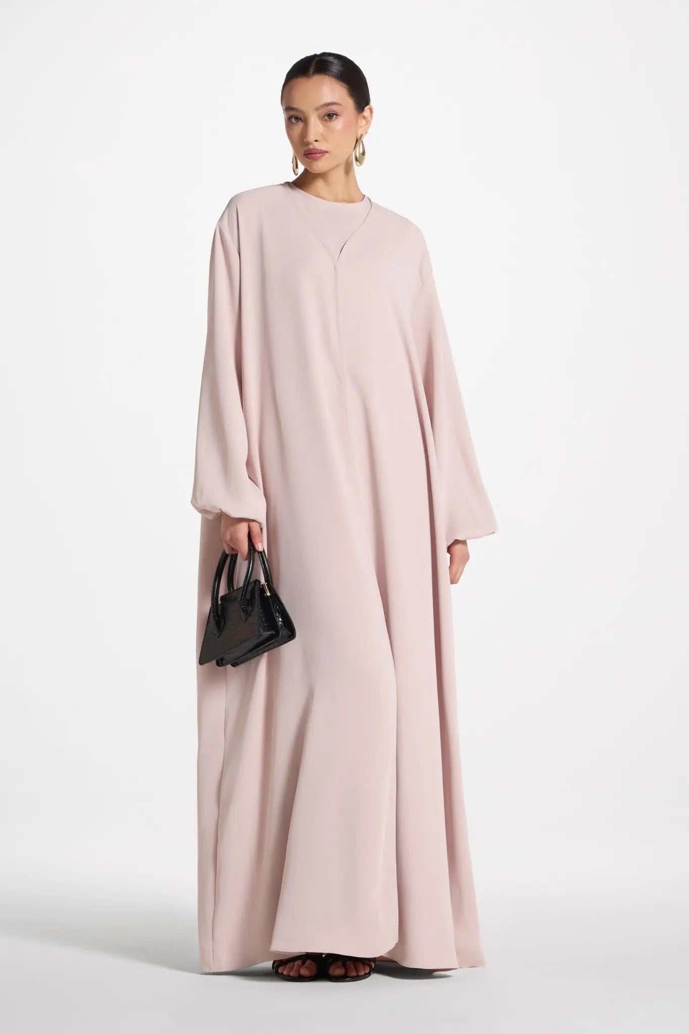 Woman wearing a long light pink dress holding a black handbag on a white background