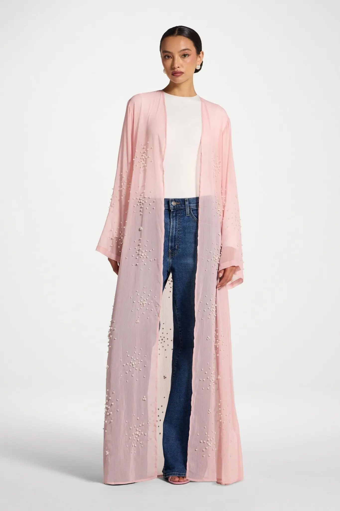 Woman wearing a long pink embroidered jacket over a white top and blue jeans on a white background