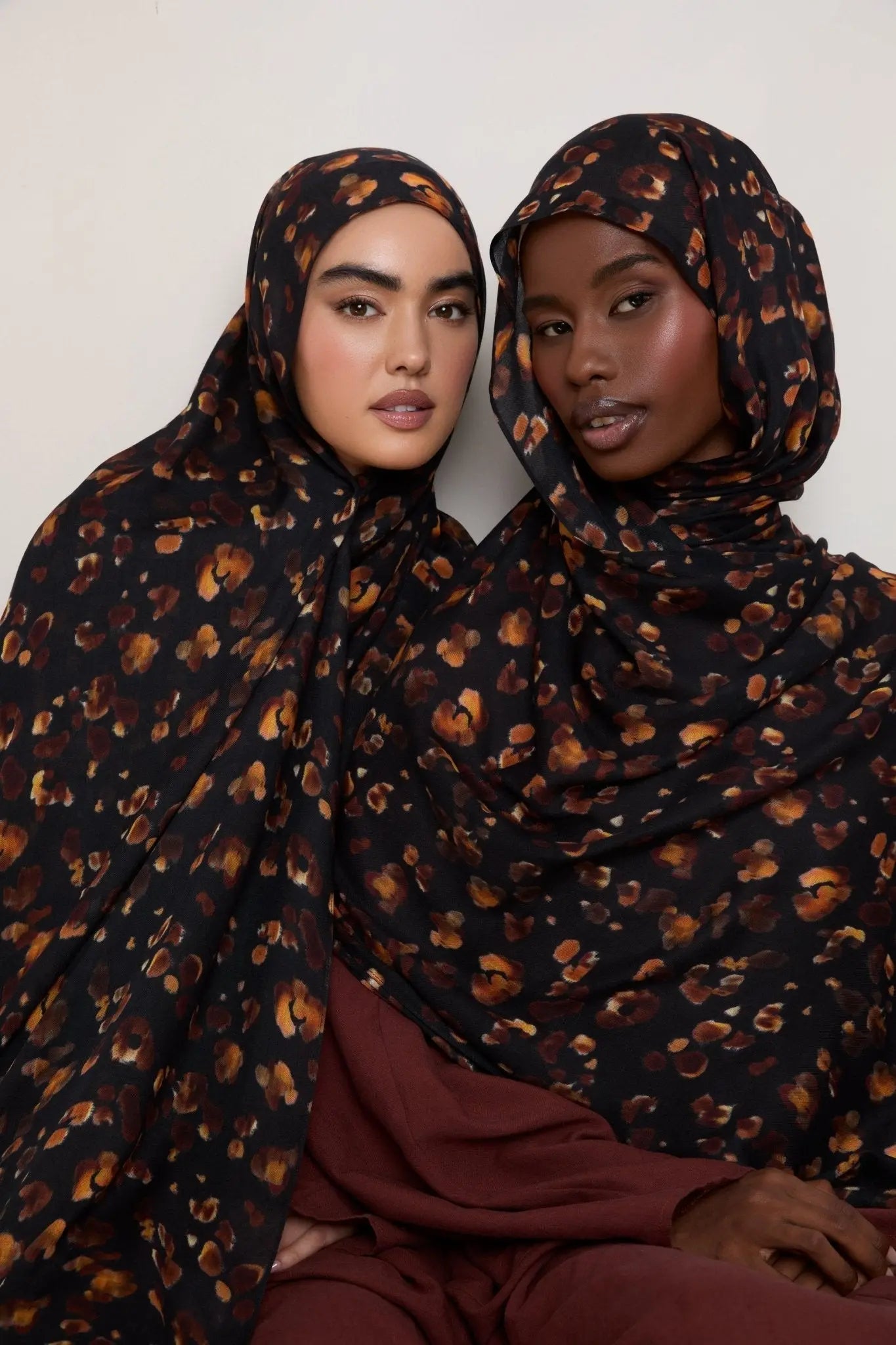 Two women wearing black hijabs with brown leopard print patterns against a white background
