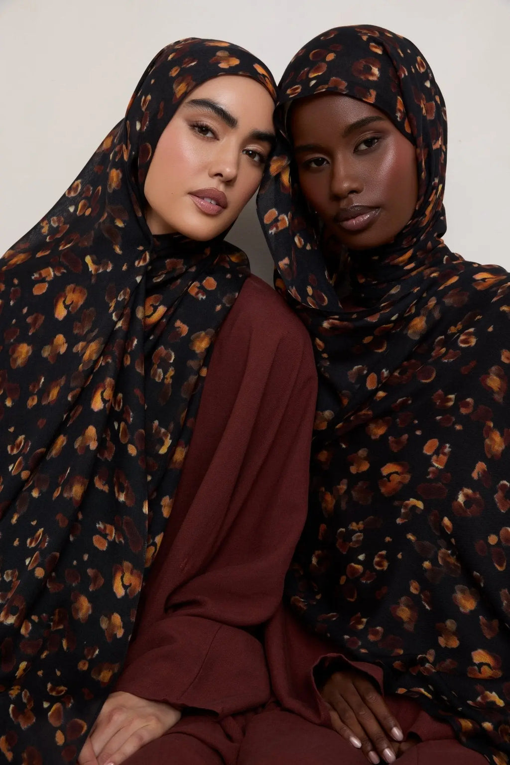 Two women wearing matching dark brown hijabs with a subtle pattern against a neutral background