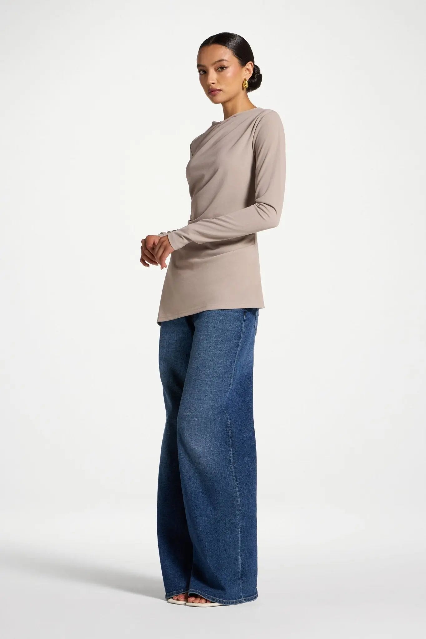 Woman wearing a beige long-sleeve top and blue jeans on a white background