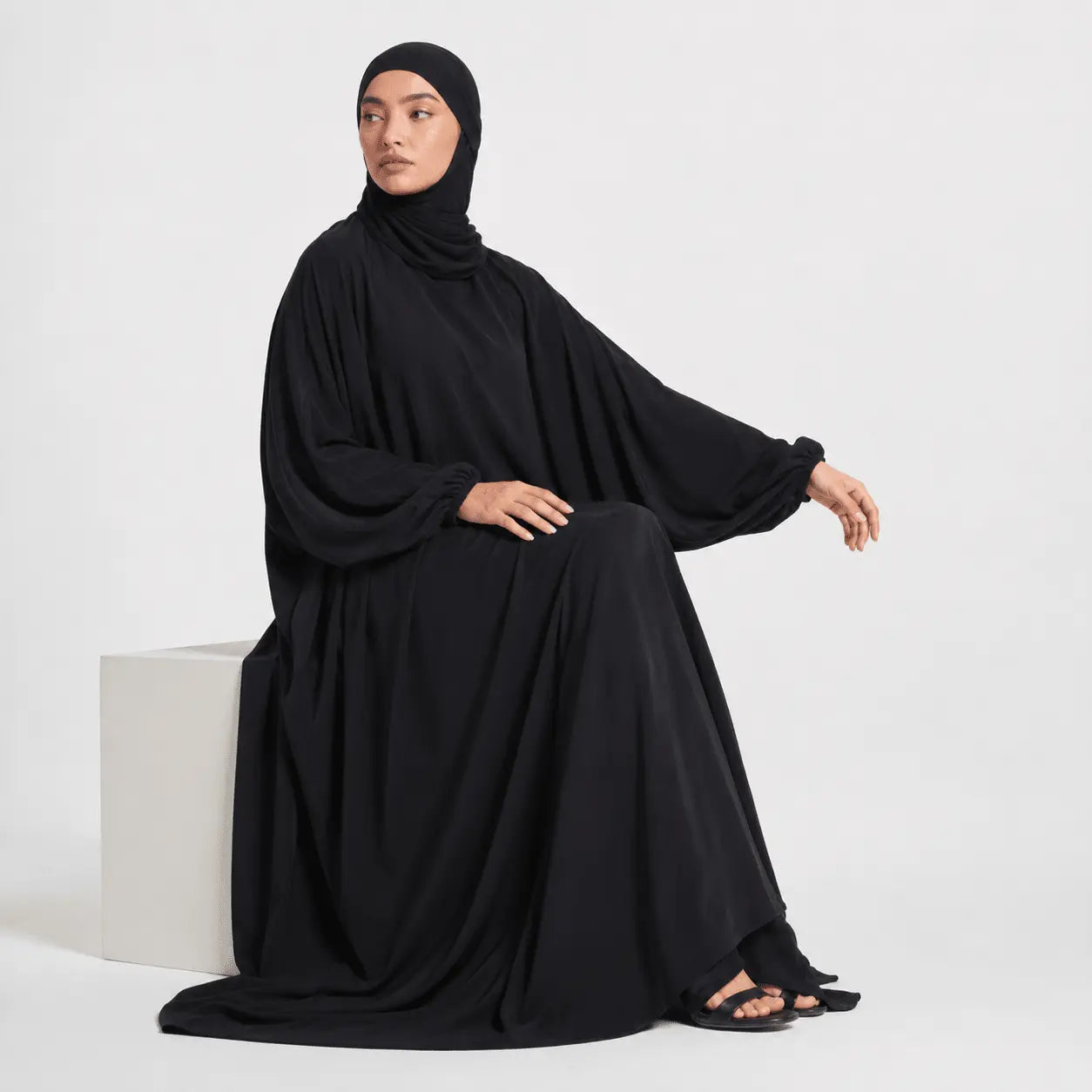 Woman wearing a black abaya sitting on a white cube against a white background