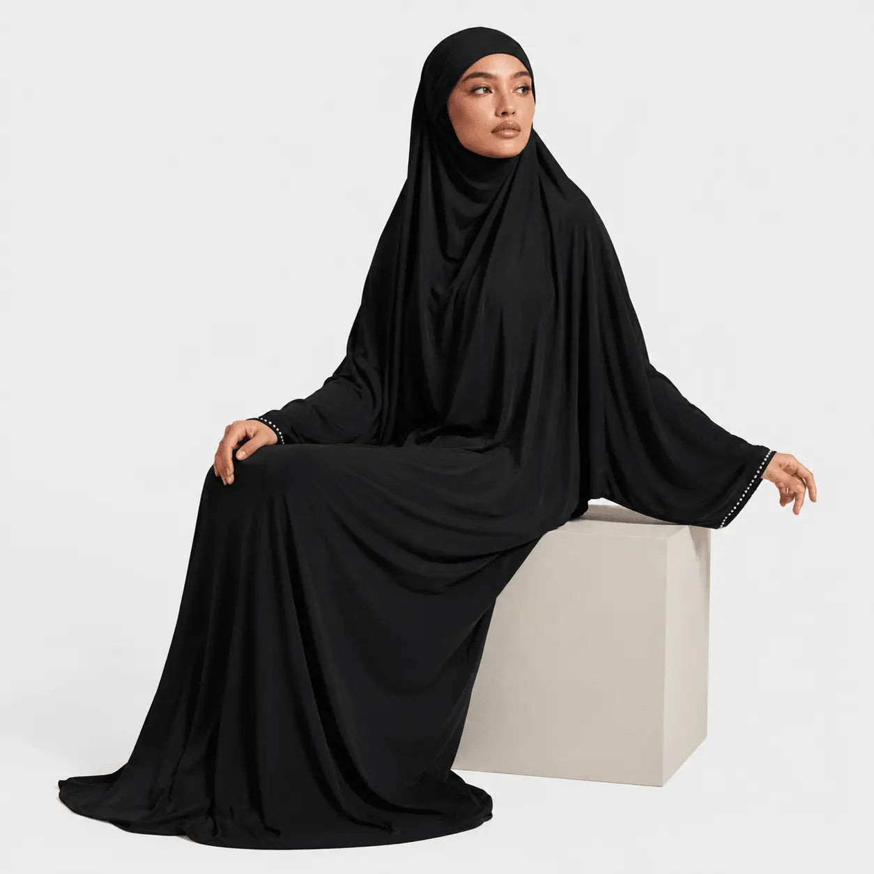 Woman wearing a black abaya sitting on a white cube against a light gray background