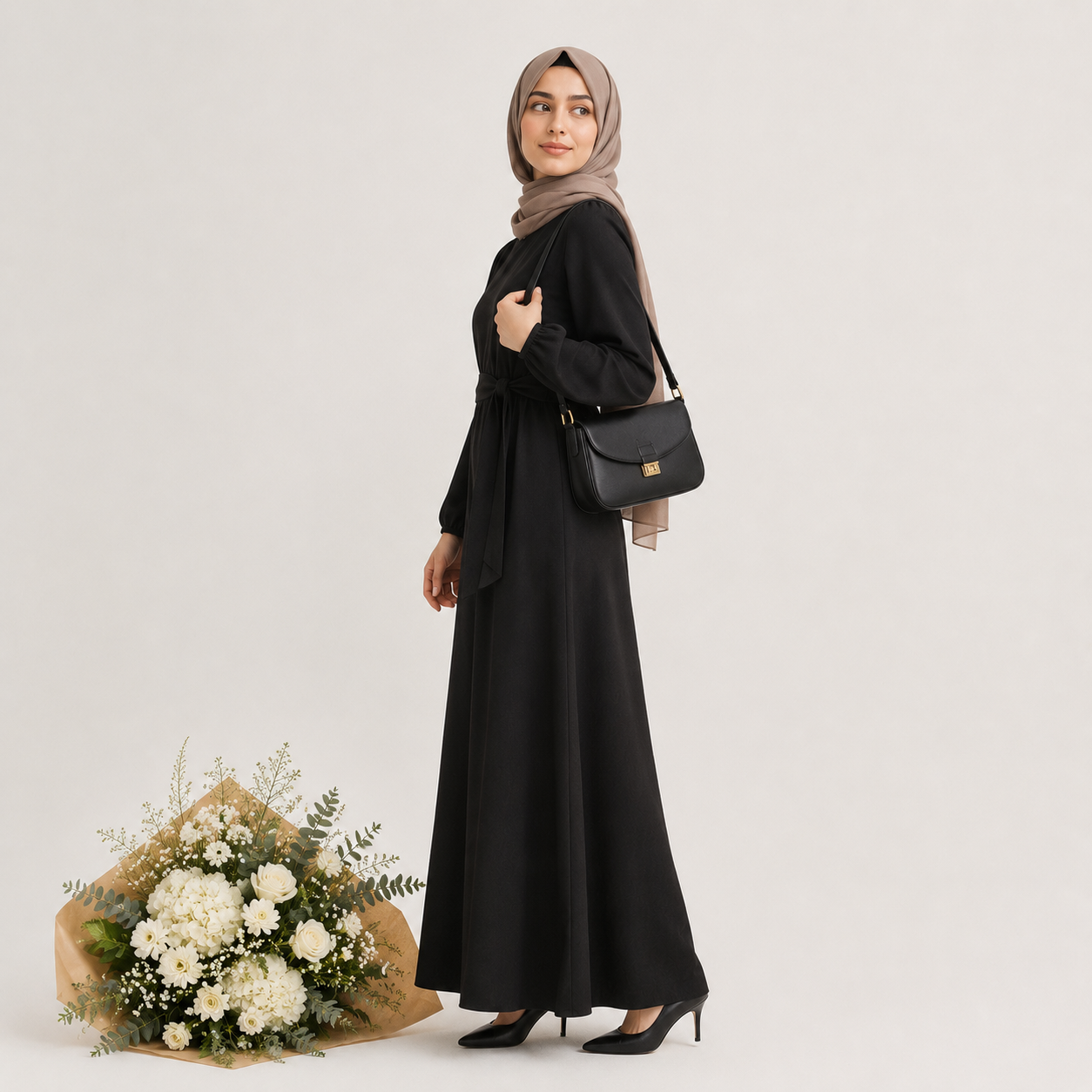 A woman wearing a long black modest dress and a taupe hijab stands in profile against a light gray studio background, carrying a black shoulder bag with a small gold logo. A bouquet of white flowers sits on the floor beside her.