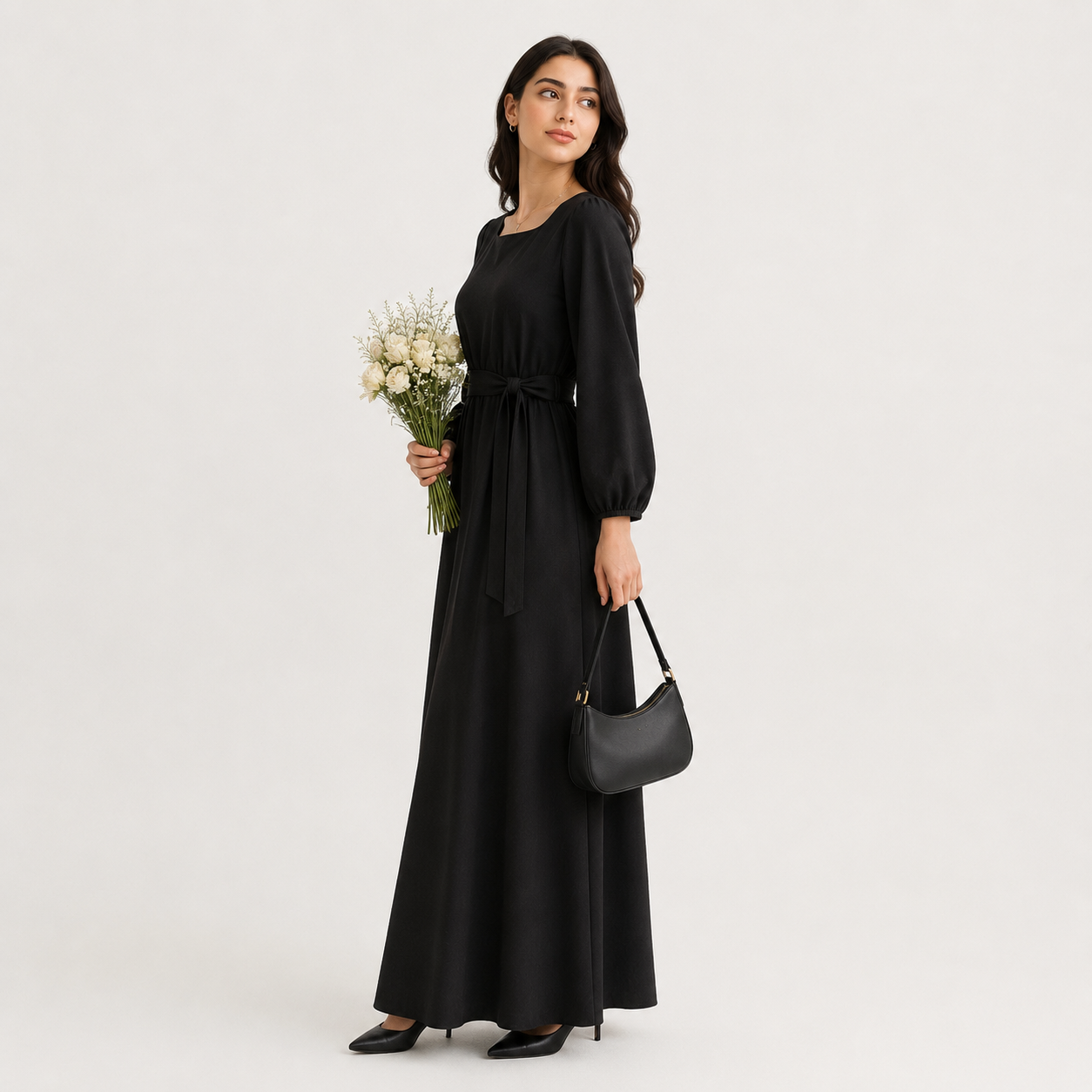 A woman modeling a long black maxi dress with a tie waist, holding a bouquet of white flowers and a black handbag against a plain light gray studio background.