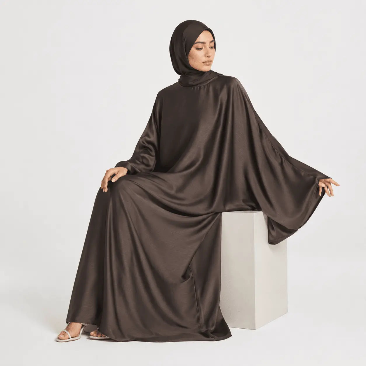Woman wearing a brown abaya sitting on a white cube against a white background