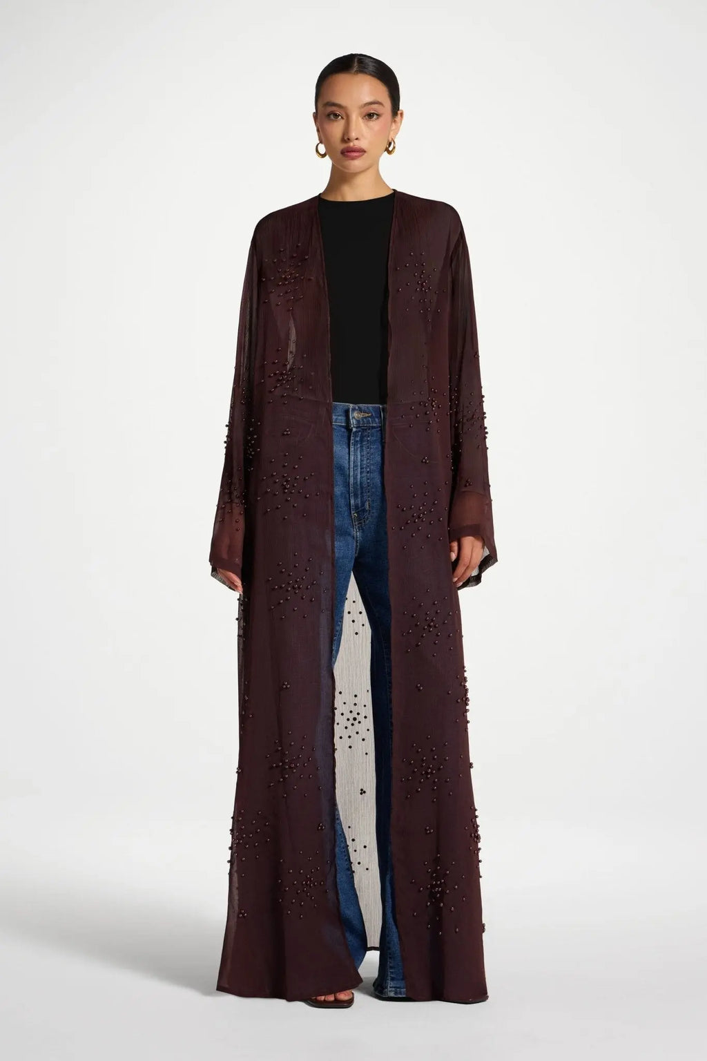 Woman wearing a long brown coat over a black top and blue jeans on a white background
