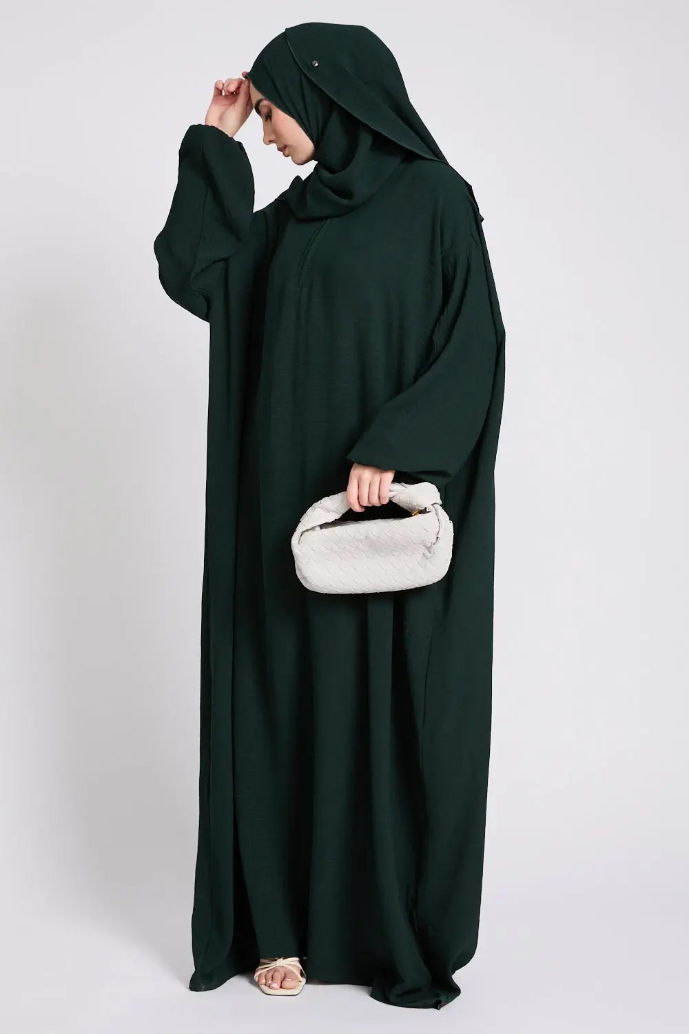 Person wearing a dark green abaya holding a white handbag on a light gray background