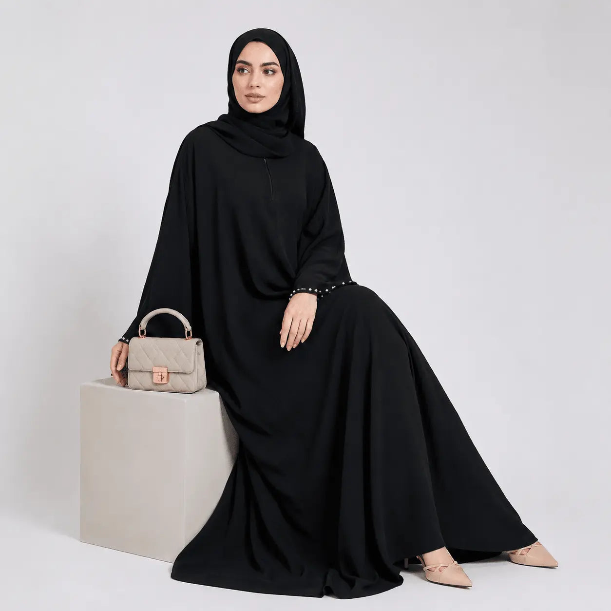 Woman in a black abaya sitting on a white box with a beige handbag.