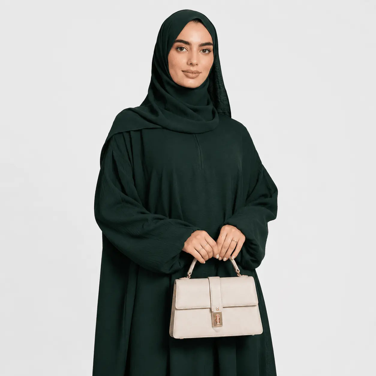 Woman in a dark green abaya holding a beige handbag against a light gray background