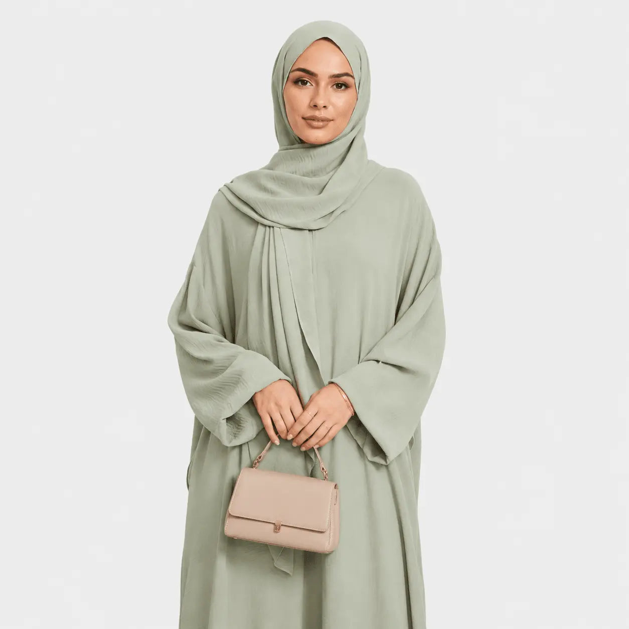 Woman wearing a light green hijab and dress holding a beige handbag on a white background