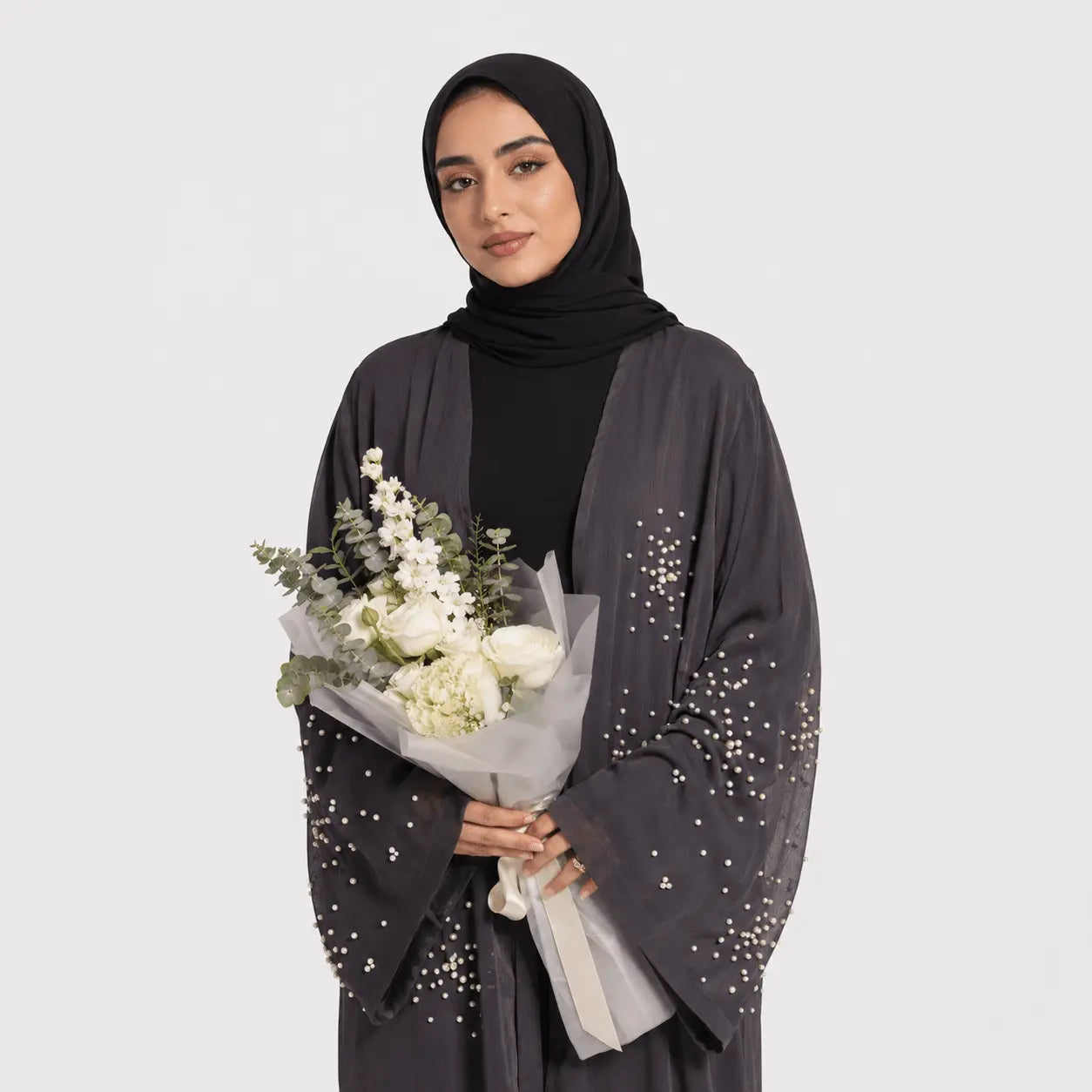 Woman holding a bouquet of flowers wearing a dark gray abaya with sequin details on a white background