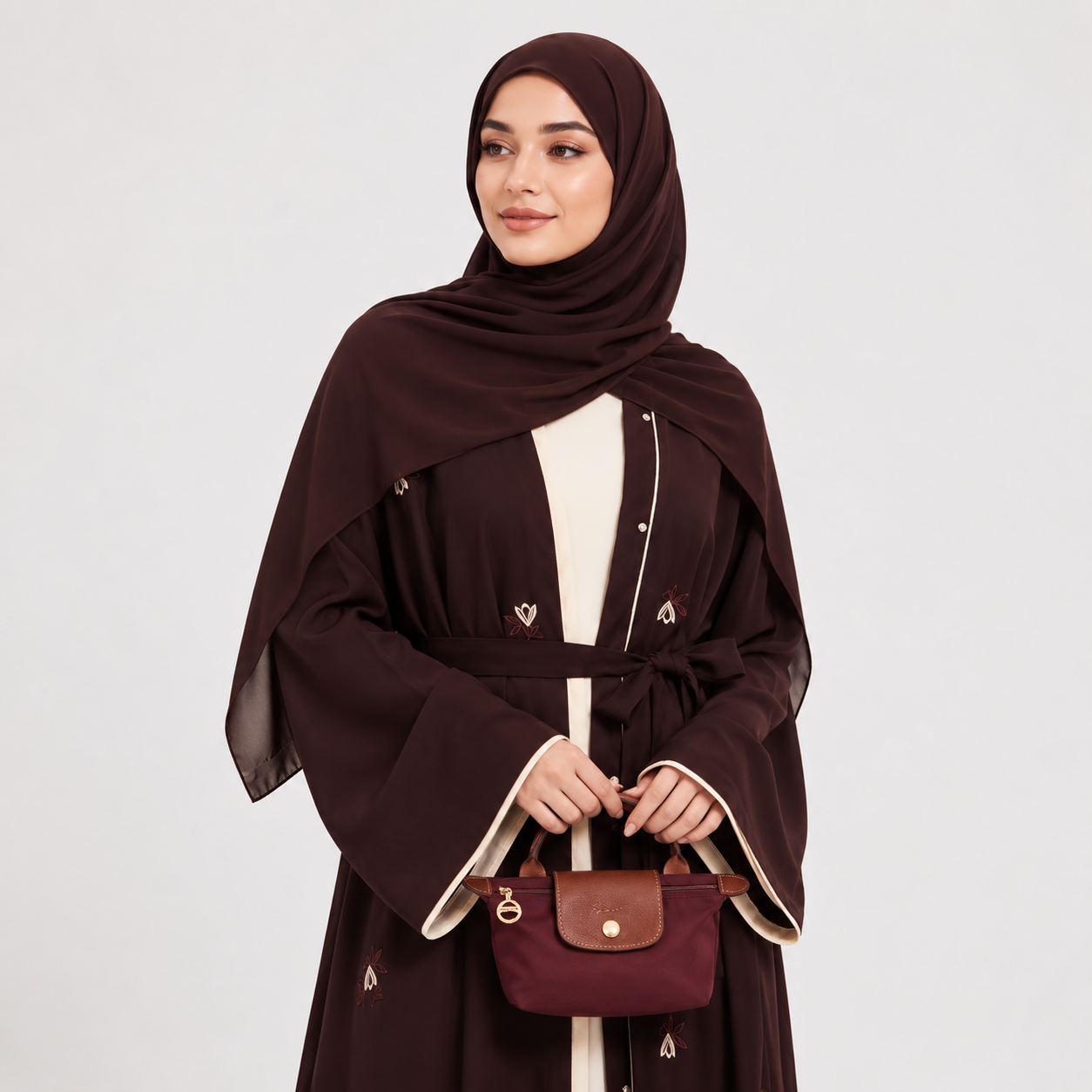 Woman wearing a dark brown abaya with a matching headscarf, holding a matching handbag on a white background.