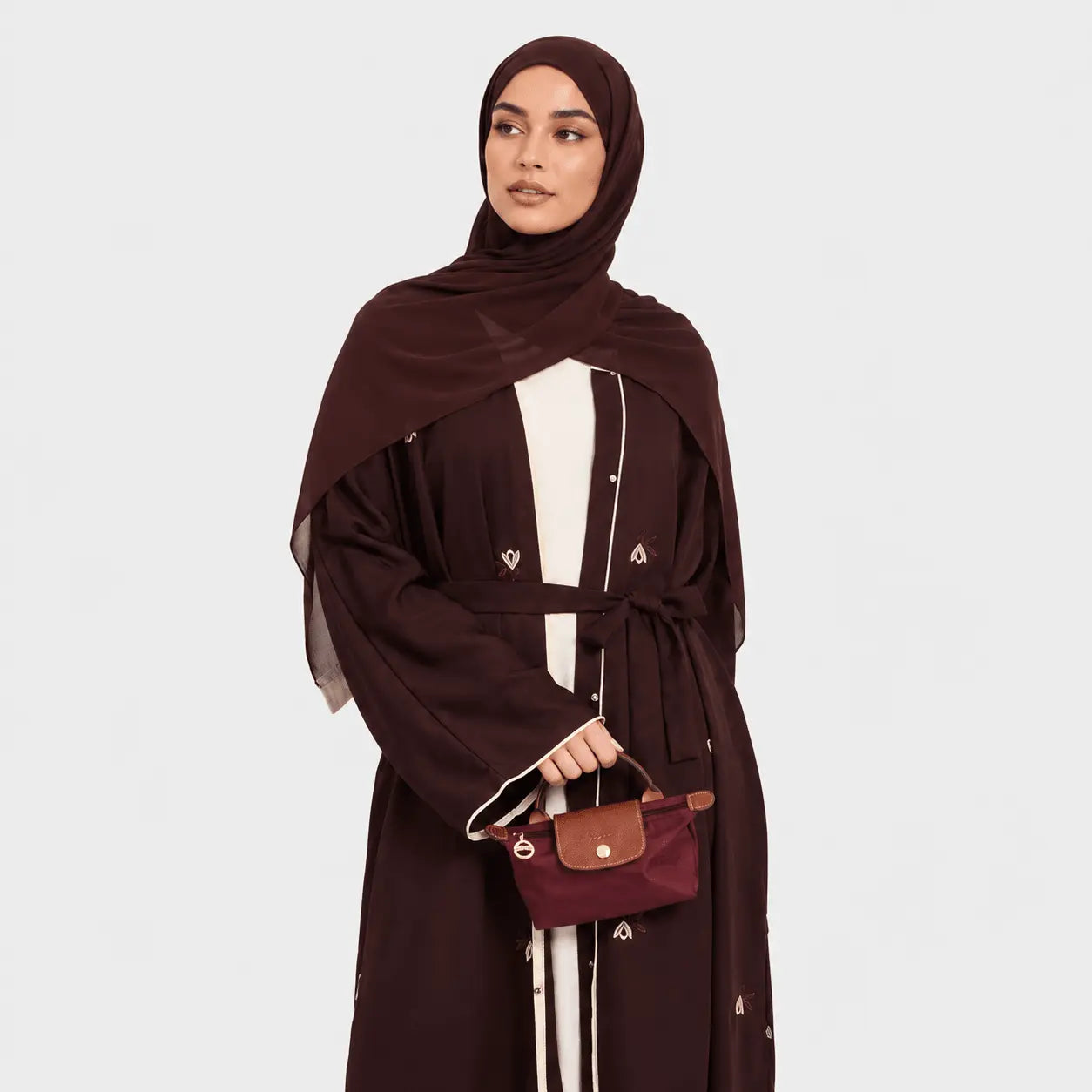 Woman wearing a brown abaya and hijab holding a matching handbag on a white background