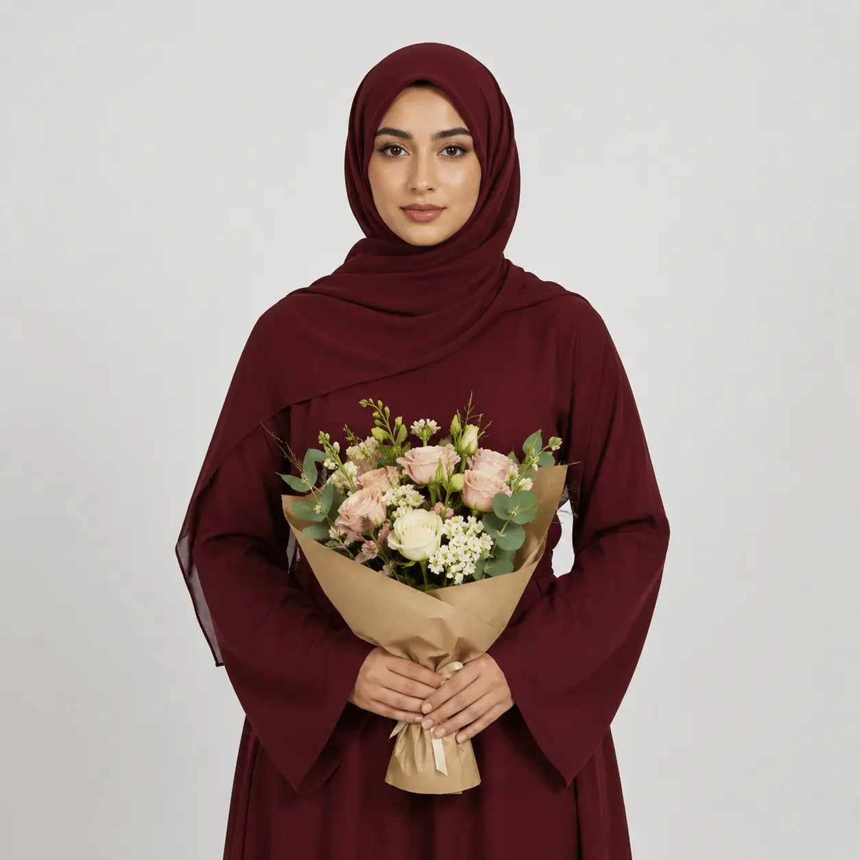 Woman wearing a burgundy hijab and dress holding a bouquet of flowers against a plain background