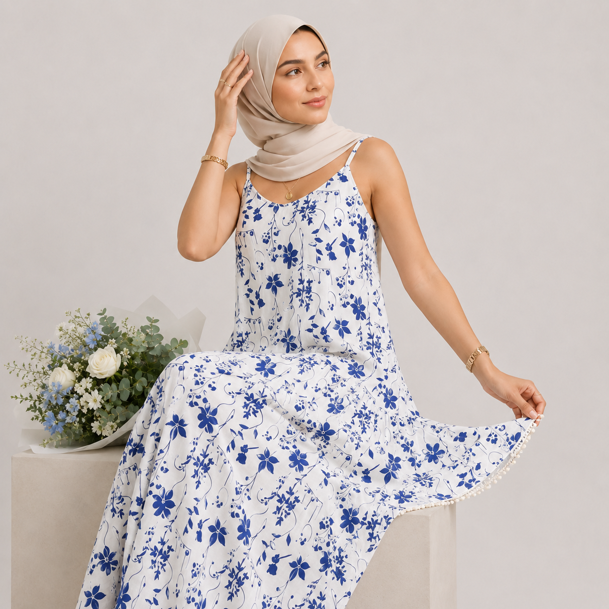 Woman wearing a white floral dress with blue flowers, sitting next to a vase of flowers on a light gray background.