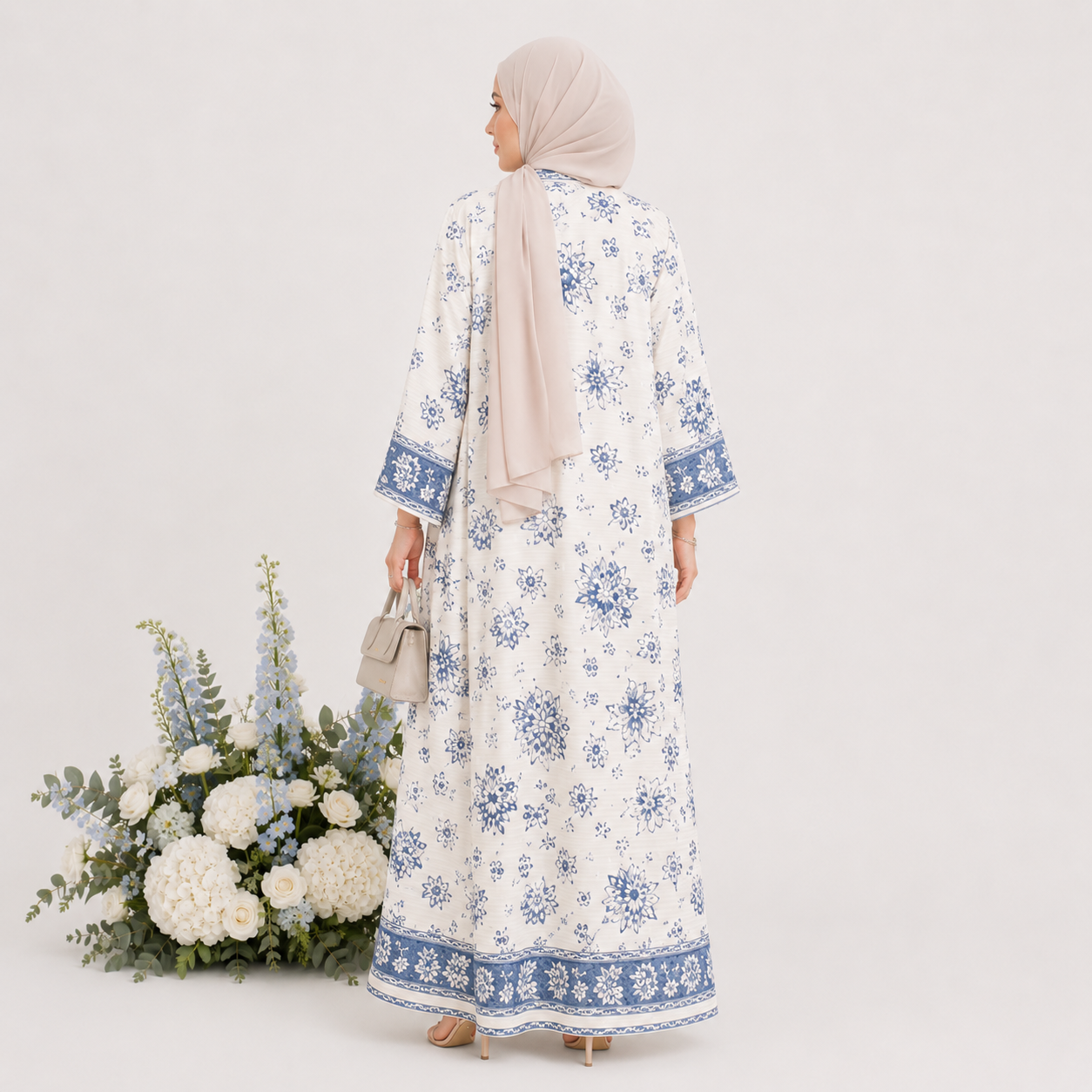 Woman wearing a white floral dress with blue details, standing next to a floral arrangement on a white background.