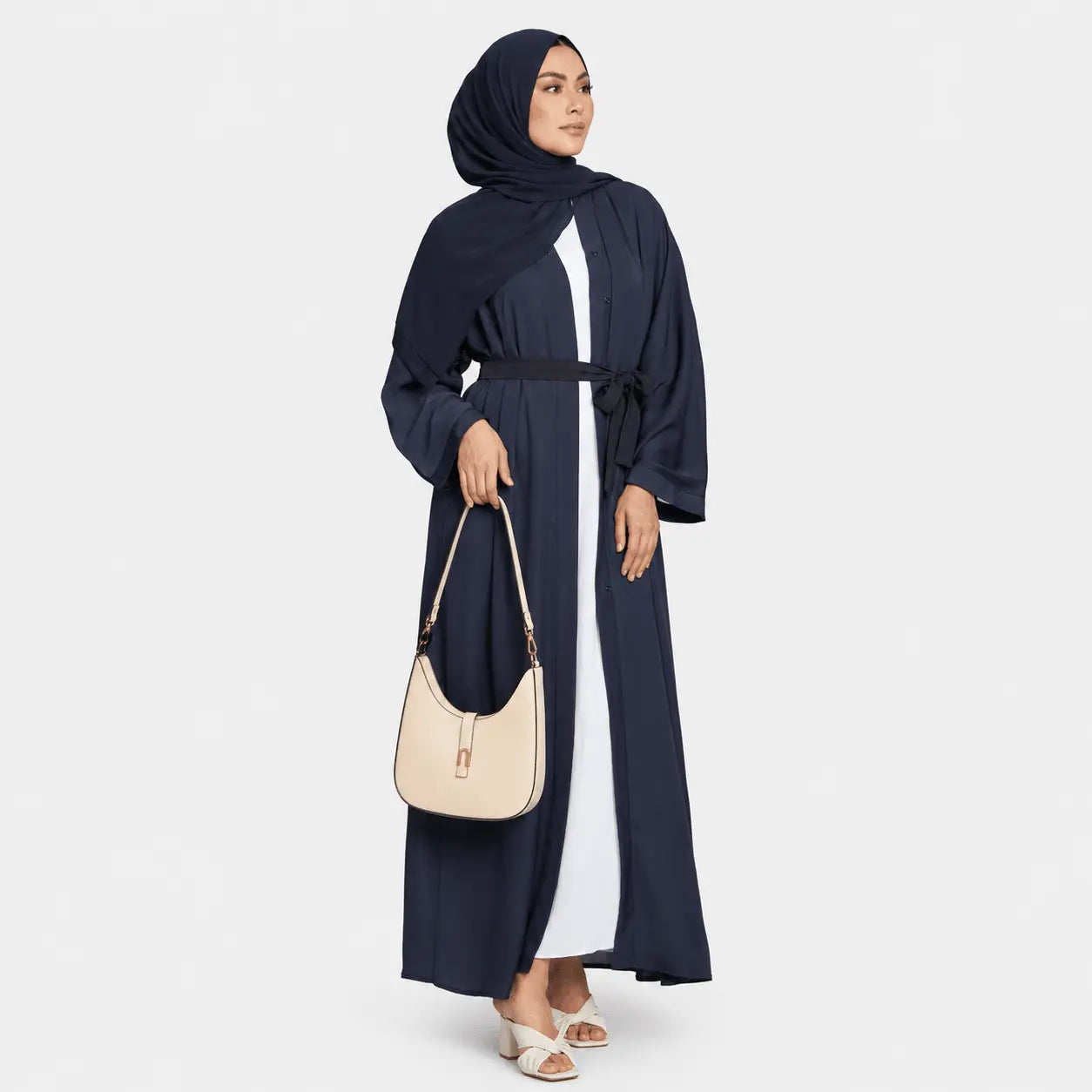 Woman wearing a navy abaya with a beige handbag on a white background