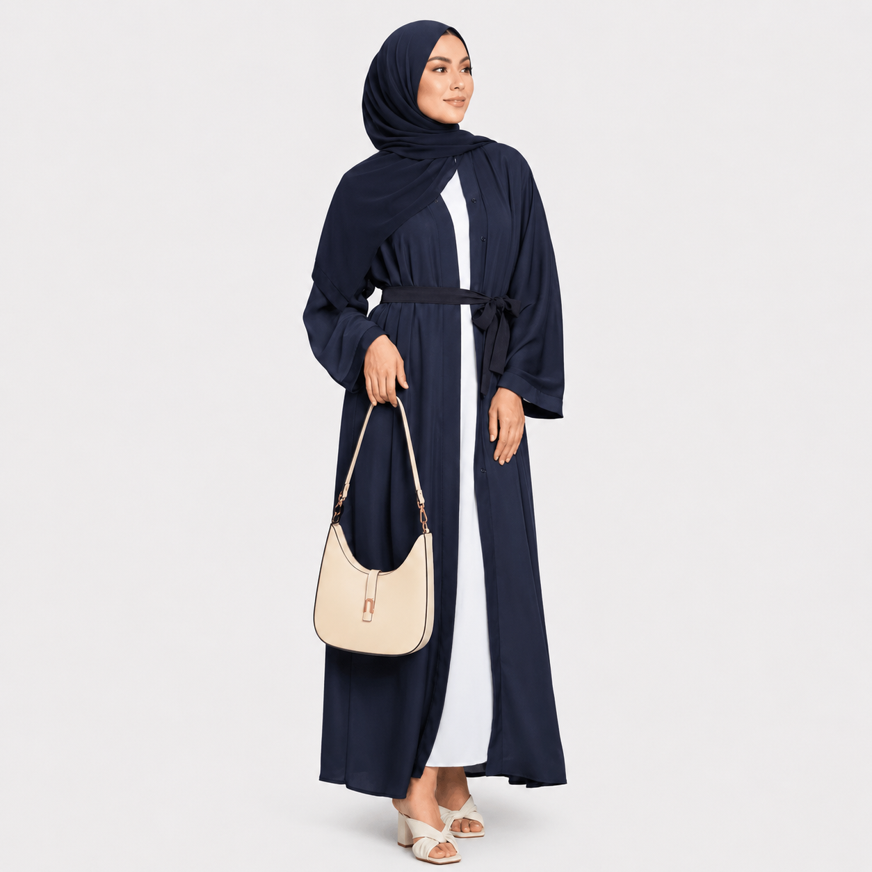 Woman wearing a navy blue abaya with a white top, holding a beige handbag on a light gray background.