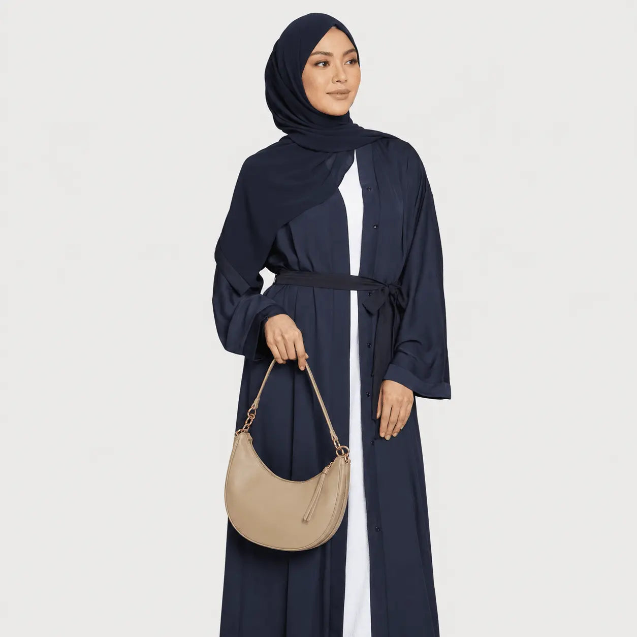 Woman wearing a navy abaya and hijab holding a beige handbag on a light gray background