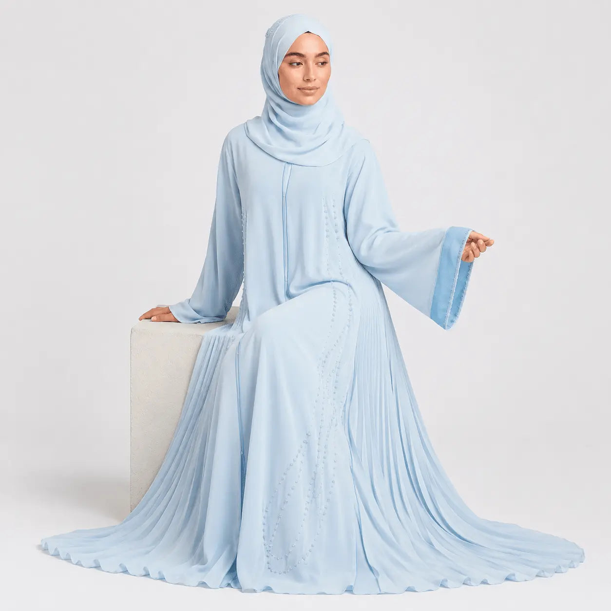 Woman wearing a light blue abaya sitting on a white cube against a white background