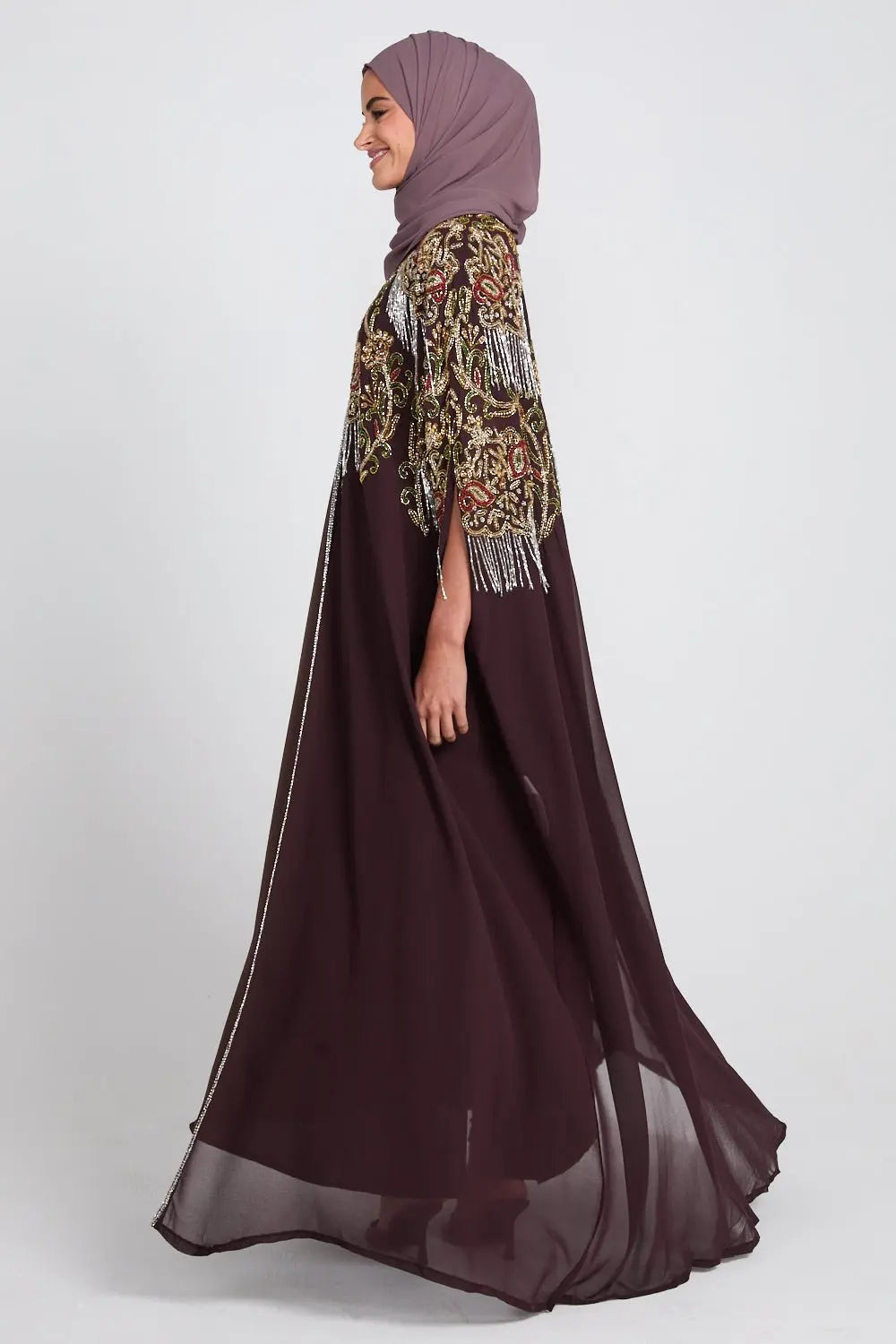 Woman wearing a long brown dress with a patterned jacket and hijab on a white background