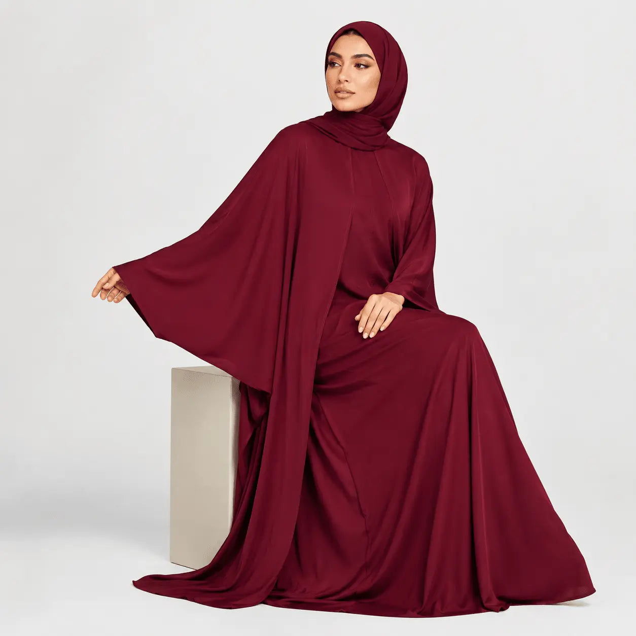 Woman wearing a burgundy abaya sitting on a white pedestal against a light gray background