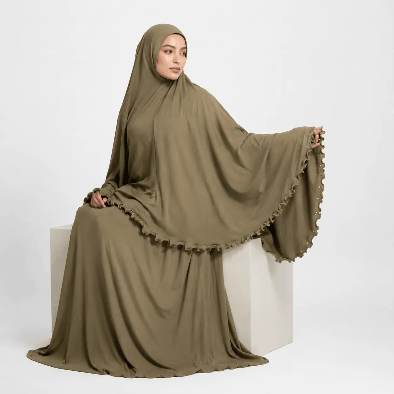 Woman wearing a brown abaya sitting on a white cube against a white background