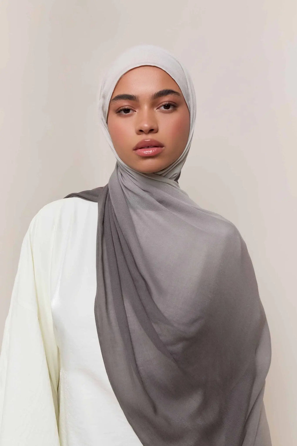 Woman wearing a gray hijab and white top against a plain background