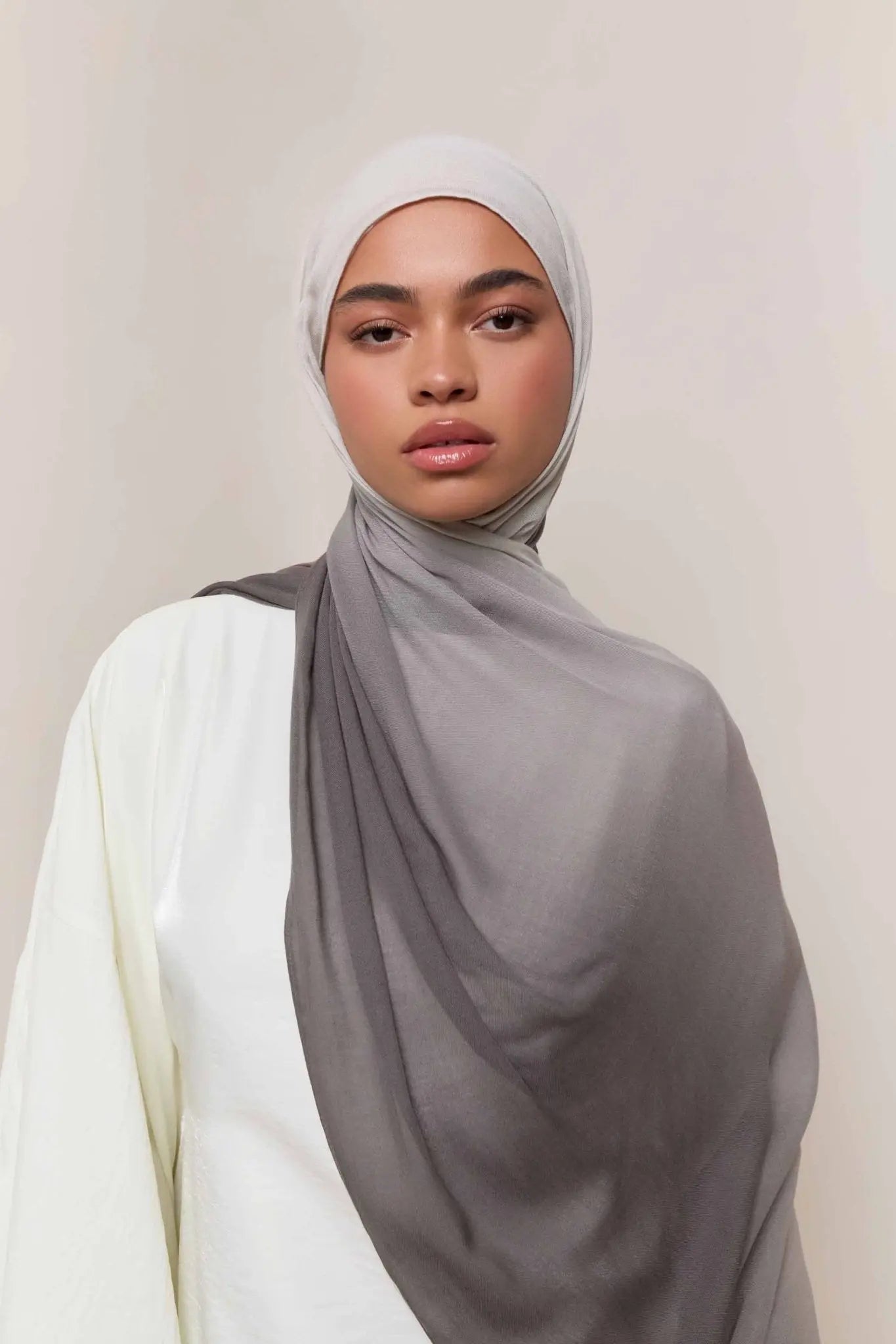 Woman wearing a gray hijab and white top against a plain background