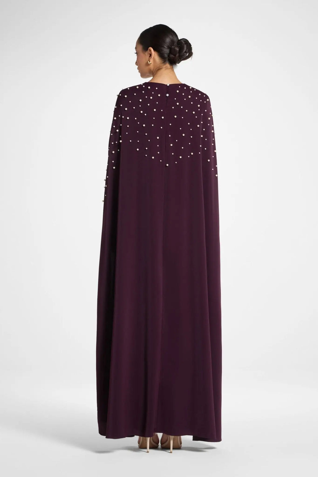Woman wearing a long, dark purple dress with a cape and embellishments on a white background