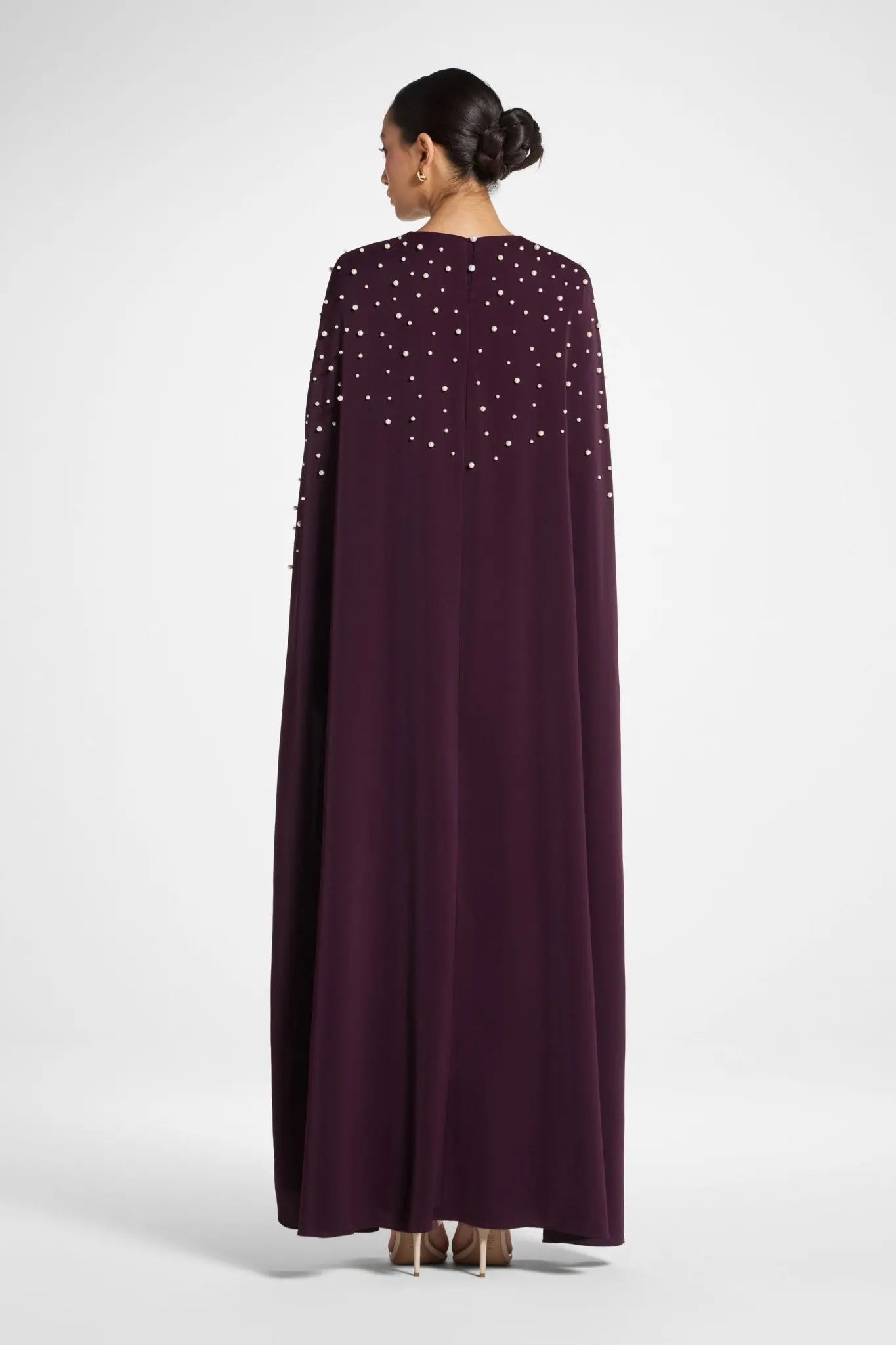 Woman wearing a long, dark purple dress with a cape and embellishments on a white background