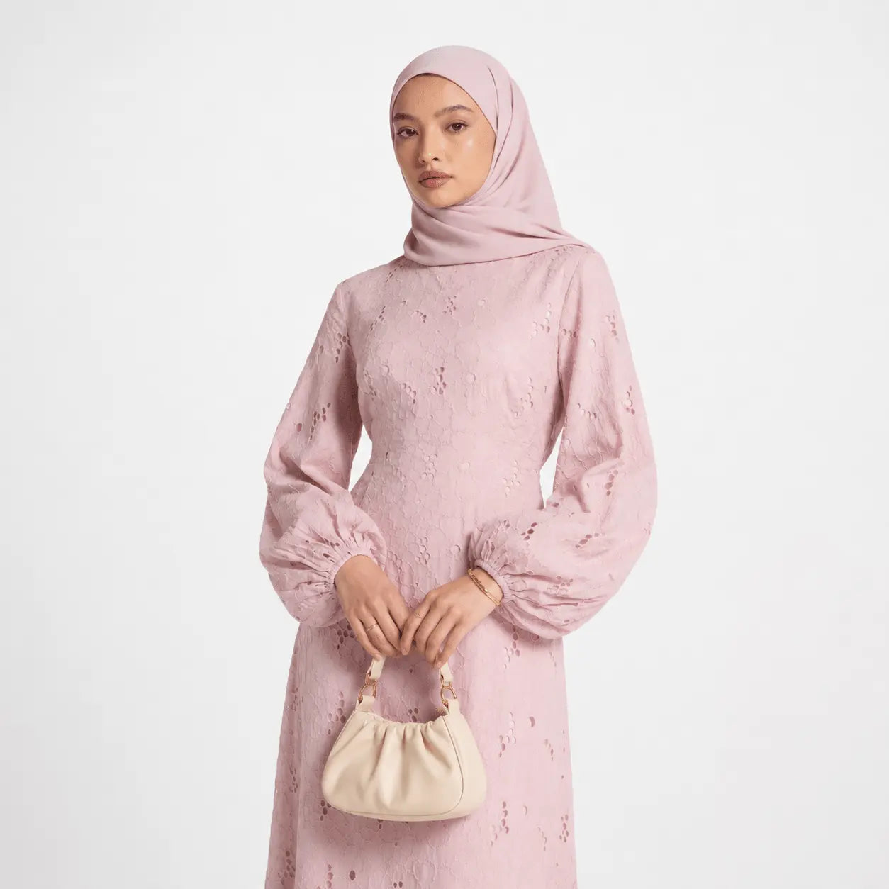 Woman in dusty pink lace dress with puff sleeves, matching hijab, holding cream handbag